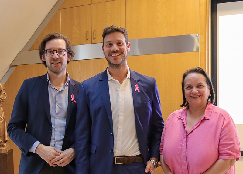 Drei Vortragende der Fortbildung „Update Brustgesundheit“ stehen nebeneinander vor holzvertäfelter Wand, zwei tragen Anzüge mit rosa Schleife, eine Frau ein pinkes Hemd.